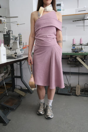 Woman standing in fashion studio wearing a mauve skirt and matching top. Paired with a neck scarf, shoulder bag and sneakers. The skirt has a crossover waistband and fits to the knees.