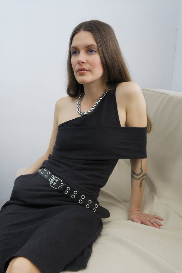 young woman sitting on a couch wearing a Black one-shoulder tank top, with a band that crosses over the chest and arm. Design by Alex Watson Studio. 