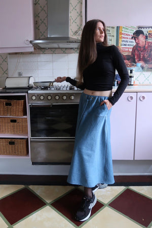 A woman with long brown hair, wearing a long-sleeve black turtleneck and a long blue denim skirt, standing in a kitchen with a casual and relaxed expression while wearing New Balance sneakers. This outfit is proudly made in Montreal.
