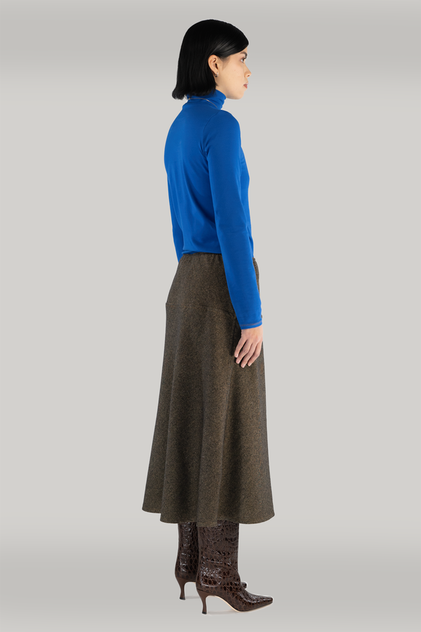 Young woman with short black hair stands confidently wearing a cobalt blue long-sleeve  turtleneck and long brown wool skirt with a bias cut. She completes her look with brown boots and gold necklace
