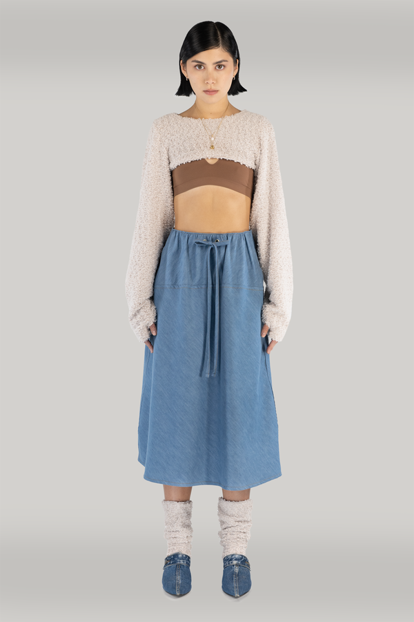 woman standing in fron of grey backdrop, wearing a beige long-sleeve sweater with thumbholws, showing brown bra and midriff. paired with a maxi denim blue skirt with drawstrings. also wearing fuzzy socks and blue heels. woman is young, fair skin with black short hair.