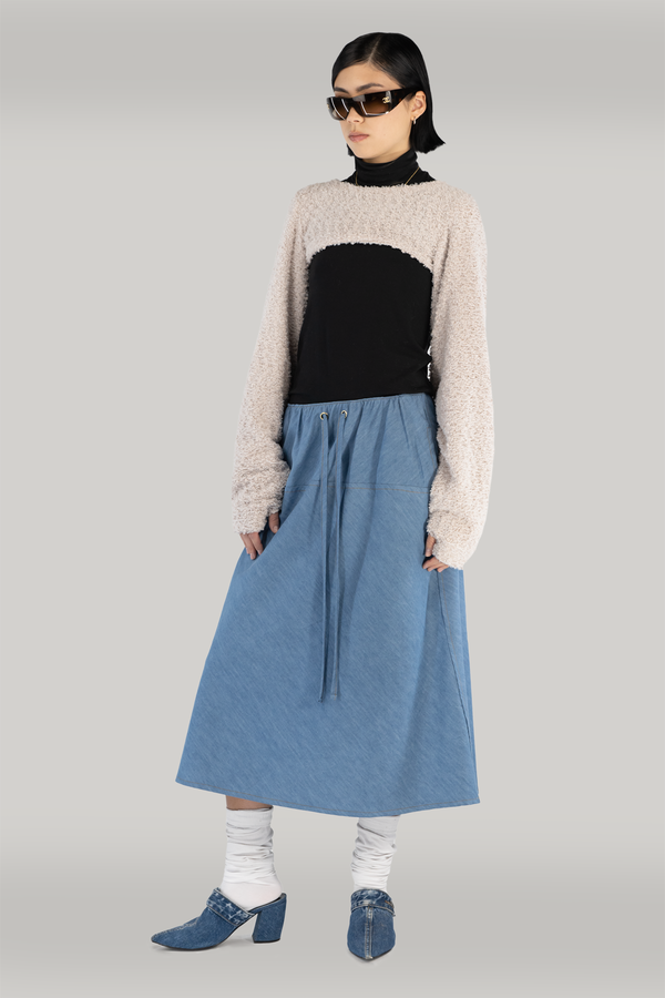 woman standing in fron of grey backdrop, wearing a beige long-sleeve sweater with thumbholws, black turtleneck paired with a maxi denim blue skirt with drawstrings. also wearing fuzzy socks and blue heels. woman is young, fair skin with black short hair.