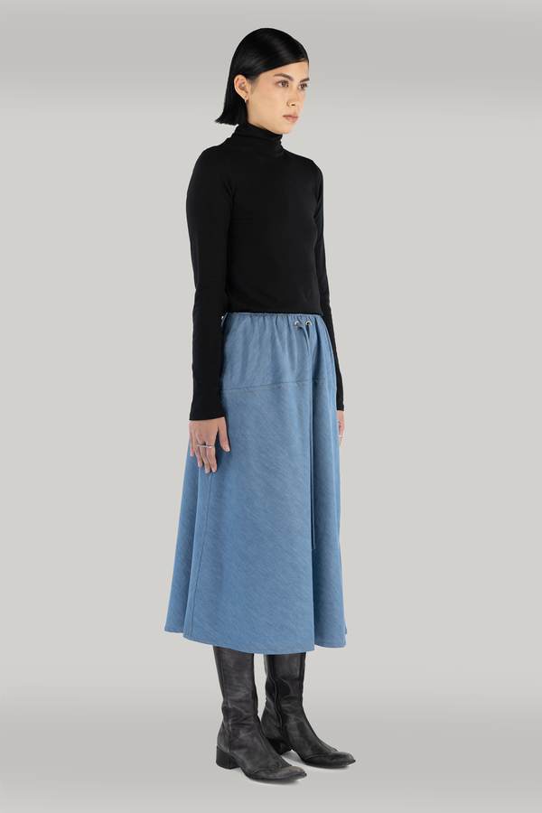 A woman with short black hair wearing a comfortable and chic long-sleeve black turtleneck and a stylish long blue denim skirt, paired with black boots. This beautifully crafted clothing is proudly made in Montreal."