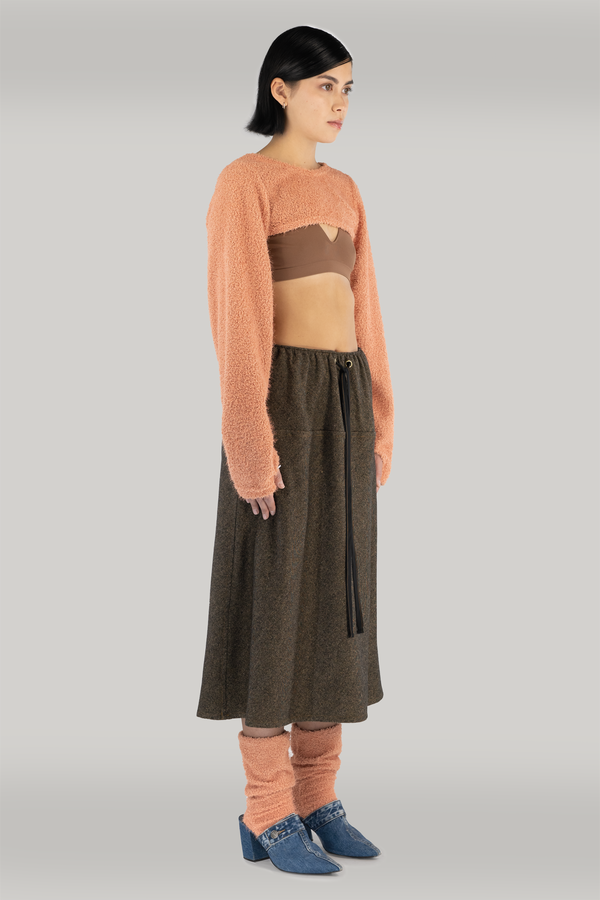 woman standing in front of grey backdrop, wearing an orange cropped shrug sweater, showing brown bra and midriff. paired with brown long skirt, orange fuzzy socks and blue heels